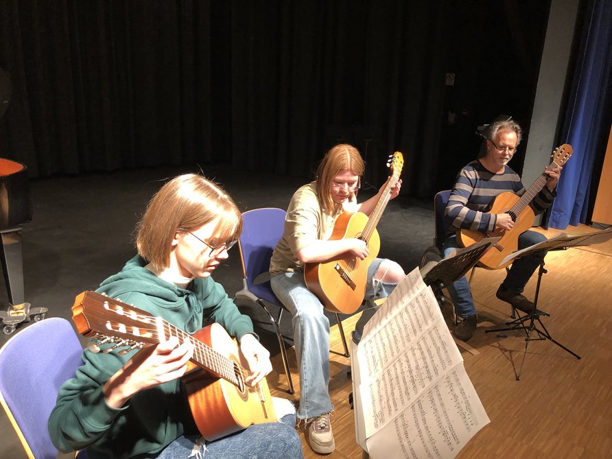 Zwei Schülerinnen mit Gitarre und ihrem Lehrer Michael Distler auf der Bühne