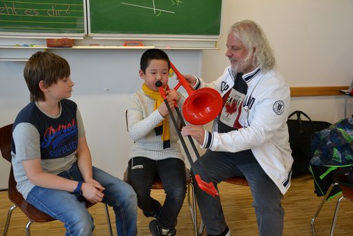 Ein Lehrer zeigt einem Jungen, wie er in eine Posaune spielen soll. Ein anderer Junge schaut zu